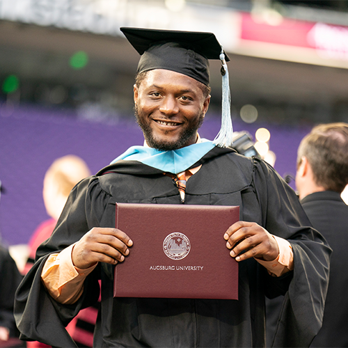 Man in graduation attire holding a diploma cover from Augsburg University.
